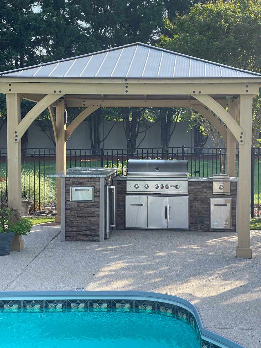 l shaped outdoor kitchen with refrigerator and side burner under wooden gazebo for coverage by a pool