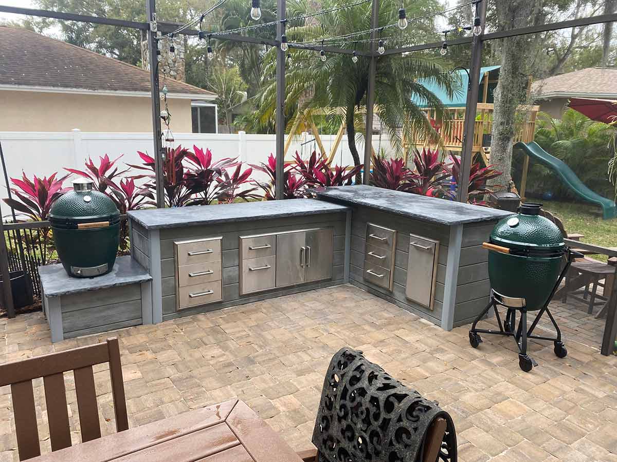 l shaped outdoor kitchen in backyard with many storage products including two 3-drawer storages access doors, and trash along with 2 asado grills