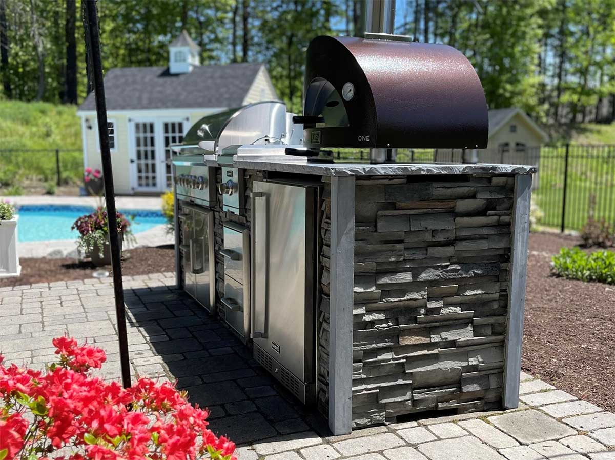 side profile of stone outdoor kitchen with pizza oven and surrounding landscaping that makes sense